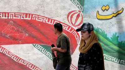 رجل وامرأة يسيران أمام لوحة جدارية تصور العلم الإيراني في أحد شوارع طهران